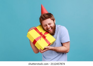 Portrait of happy positive handsome bearded man in party cone embracing yellow gift box, getting present from friend on his birthday. Indoor studio shot isolated on blue background.