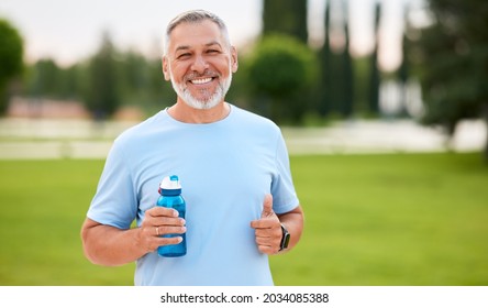 Portrait Of Happy Positive Mature Man With Broad Smile Holding Water Bottle While Doing Sport In City Park, Active Retired Male Sportsman Jogging Outside In Early Morning. Healthy Lifestyle Concept