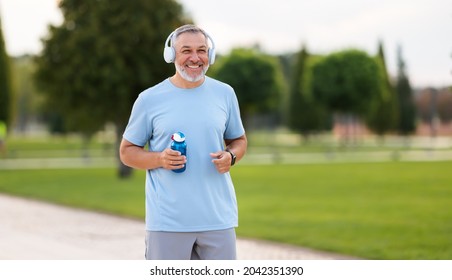 Portrait Of Happy Positive Mature Man With Broad Smile Holding Water Bottle While Doing Sport In City Park, Active Retired Male Sportsman Jogging Outside In Early Morning. Healthy Lifestyle Concept