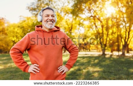 Portrait of happy positive mature man with broad smile  in headphones while doing sport in city park, active retired male sportsman jogging outside in early morning. Healthy lifestyle concept