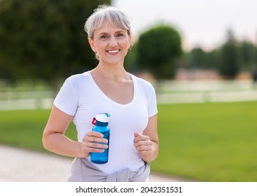 Portrait Of Happy Positive Mature Woman With Broad Smile Holding Water Bottle While Doing Sport In City Park, Active Retired Female Sportsman Jogging Outside In Early Morning. Healthy Lifestyle Concep