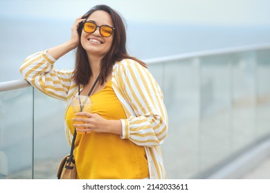 Portrait Of Happy Positive Millennial Plus Size Woman 30-35 Years Old On Vacation. Smiling Overweight Female In In Yellow And White Fashion Clothes And Sunglasses Drinks Juicy Coctail In Summer Day