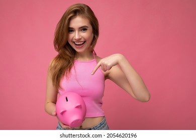 Portrait Of Happy Positive Smiling Young Pretty Attractive Woman With Light Hair Wearing Stylish Pink Top Isolated Over Pink Background With Empty Space And Holding Pink Pig Penny Bank. Savings Box