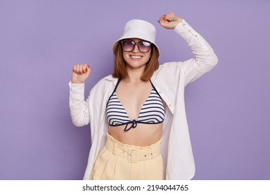 Portrait Of Happy Positive Woman Wearing Panama, White Shirt And Swimming Suit Posing Isolated Over Purple Background, Standing With Raised Arms, Rejoicing Her Success.