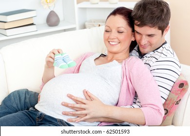 Portrait of a happy pregnant woman holding baby shoes and of her husband on a sofa at home