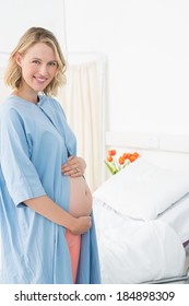 Portrait Of Happy Pregnant Woman Wearing Gown Standing By Bed In Hospital Ward