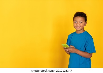 Portrait Of Happy Preteen Boy Holding Mobile Phone. Multiracial Child Wearing Blue T-shirt Looking At Camera And Smiling Against Yellow Background. Mobile Technology Concept