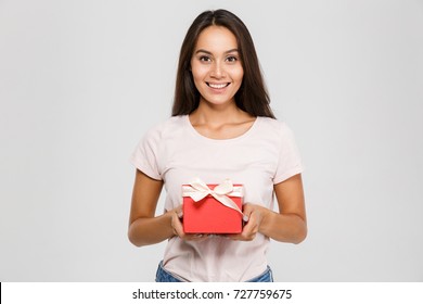 Portrait of a happy pretty asian woman holding present box while standing and looking at camera isolated over white background