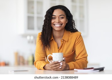 Portrait of happy pretty long-haired young black woman in yellow shirt sitting at kitchen table with cup of herbal tea and smiling at camera, enjoying morning coffee at home, copy space