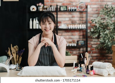 Portrait of happy professional nail master sitting at working place in salon. young beautiful asian japanese lady manicurist worker in nail workshop. beautician in apron face camera smiling in studio - Powered by Shutterstock