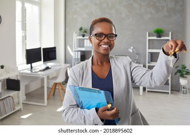 Portrait of happy real estate agent or mortgage broker in suit and glasses showing key to new house. Beautiful short haired young black woman standing in office, holding clipboard and keys and smiling