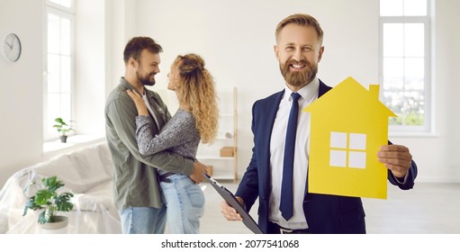 Portrait Of Happy Real Estate Agent, Realtor Or Mortgage Broker In Suit Holding Clipboard And Home Symbol, Looking At Camera And Smiling Standing In Living Room Of New House After Meeting With Clients