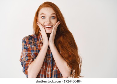 Portrait of happy redhead girl hear amazing news, looking surprised and happy, touching cheeks and smiling at camera, white background.
