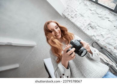 Portrait of happy redhead girl smiling and holding professional digital camera, Captivating image of a young professional woman posing with camera gear in a well-equipped studio illuminated by lights.
