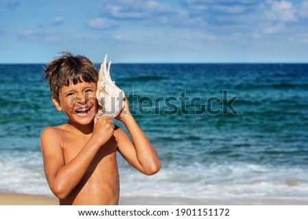 Portrait of a happy relaxed boy listen to huge seashell with big smile over sea background