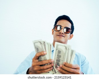 Portrait Of Happy, Rich Asian Young Businessman In Casual Blue Shirt And Wearing Sunglasses Enjoying While Holding And Counting Dollars Money Banknotes On White Wall Background.