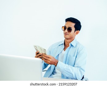 Portrait Of Happy, Rich Asian Young Businessman In Casual Blue Shirt And Wearing Sunglasses Enjoying While Holding And Counting Dollars Money Banknotes On White Wall Background.