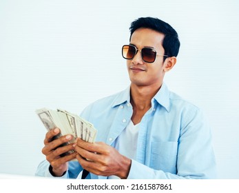 Portrait Of Happy, Rich Asian Young Businessman In Casual Blue Shirt And Wearing Sunglasses Enjoying While Holding And Counting Dollars Money Banknotes On White Wall Background.