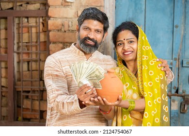 Portrait Of Happy Rural Couple Holding Indian Rupee Notes And Clay Money Box Or Gullak. Traditional Piggy Bank, Save Money, Investment And Banking