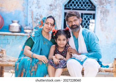 Portrait Of Happy Rural Indian Parents With Girl Child, Family Sitting Outside Village Home, Cute Adorable Little Daughter With Her Father And Mother.  Man Wear Traditional Kurta And Woman Wear Saree 