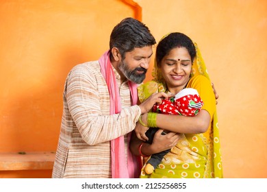 Portrait Of Happy Rural Indian Parents With Newborn Baby, Couple Father Mother Holding New Born Baby Against Orange Background.