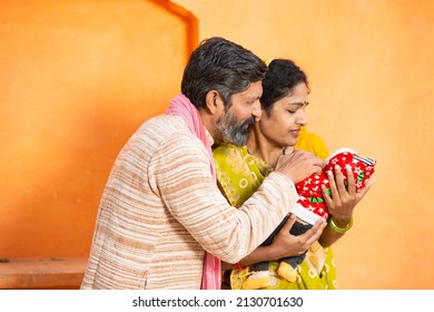 Portrait Of Happy Rural Indian Parents With Newborn Baby, Couple Father Mother Holding New Born Baby Against Orange Background.