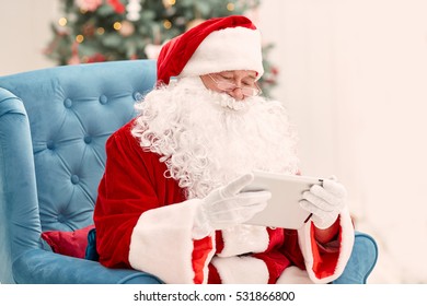 Portrait of happy Santa Claus holding Christmas letter using tablet on the armchair at home in the living room - Powered by Shutterstock