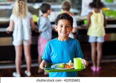 Portrait Of Happy Schoolboy Holding Food Tray In Canteen Against Classmates