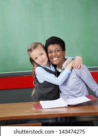 Portrait of happy schoolgirl hugging female professor at desk in classroom