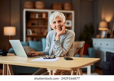 A portrait of a happy senior adult woman leaning on a desk with her chin in her hand while using a laptop and working from home. - Powered by Shutterstock