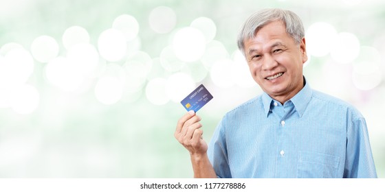 Portrait Of Happy Senior Asian Man Holding Credit Card And Showing On Hand Smiling And Looking At Camera On Isolated Blurred Bokeh Background Banner Feeling Positive Or Enjoy. Older Lifestyle Concept.