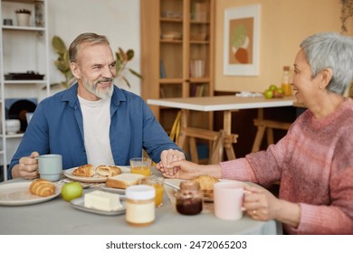 Portrait of happy senior couple holding hands at dinner table and enjoying breakfast together at home - Powered by Shutterstock