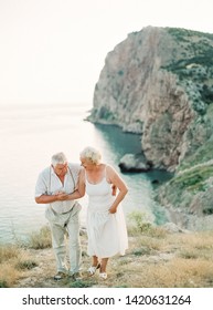 Portrait Of Happy Senior Couple On Nature. Happy Older People Walking And Smiling In Summer Near Sea Coast During Sunset. Lifestyle Elderly Retirement Concept.