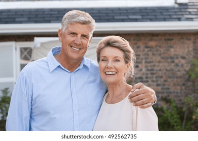 Portrait Of A Happy Senior Couple Smiling In Front Of Their House. Older Couple Embracing And Looking At Camera. Happy Old Retired Couple Hugging.