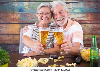 Portrait of happy senior couple toasting with beers sitting at wooden table, looking at camera smiling