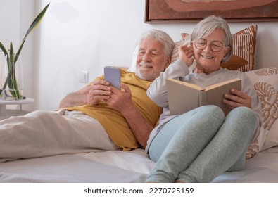 Portrait of happy senior couple together in bed, woman reading book, man using phone - Powered by Shutterstock