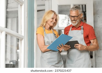 Portrait Of Happy Senior Farmers Couple With Clipboard And Digital Tablet, Smiling Older Man And Woman Wearing Aprons Checking List Of Duties, Standing At Terrace Of Their Greenhouse, Free Space - Powered by Shutterstock