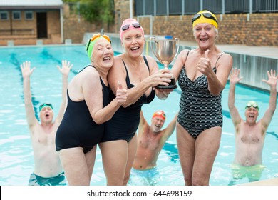 Portrait Of Happy Senior Female Swimmers Holding Trophy At Poolside