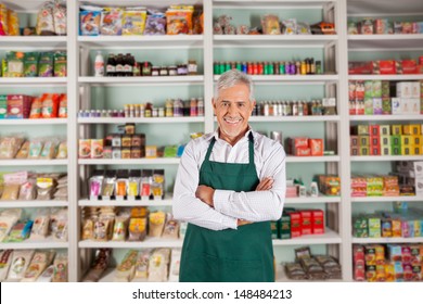 Portrait of happy senior male owner standing arms crossed against shelves in supermarket