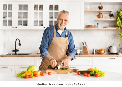 Portrait of happy senior man cooking food in kitchen at home, handsome elderly gentleman preparing lunch or dinner at home, older male wearing apron chopping vegetables and smiling at camera - Powered by Shutterstock