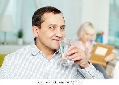 Portrait Of Happy Senior Man Drinking Water From Glass And His Wife Reading Book Behind