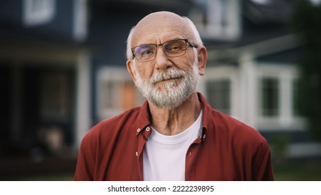 Portrait of a Happy Senior Man with Gray Hair Wearing Glasses and a Red Shirt Standing Outside in Front of a Suburbs Area House. Old Adult Man Posing and Looking at Camera and Smiling.