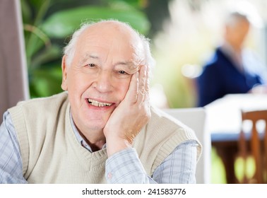 Portrait Of Happy Senior Man Sitting At Nursing Home With Grandson In Background