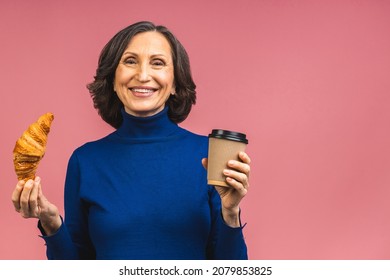 Portrait Of A Happy Senior Mature Aged Woman Eating Croissant And Drinking Coffee Isolated Over Pink Background. French Breakfast Concept.