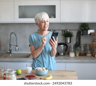 Portrait of a happy senior mature mid aged woman having a healthy breakfast after fitness exercise training and using a smartphone, looking at phone in the morning at home - Powered by Shutterstock