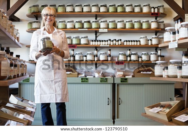Portrait of a happy senior merchant standing with\
spice jar in store