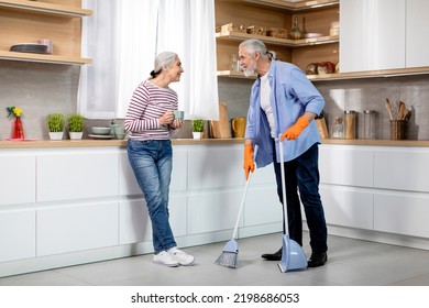 Portrait Of Happy Senior Spouses Making Cleaning In Kitchen Together, Smiling Elderly Couple Sharing Domestic Chores, Woman Drinking Tea While Her Husband Mopping Floor At Home, Copy Space