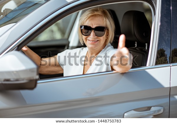 Portrait of a happy senior woman driver looking out\
the car window and showing ok sign. Concept of an active people\
during retirement age