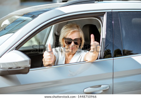 Portrait of a happy senior woman driver looking out
the car window and showing ok sign. Concept of an active people
during retirement age