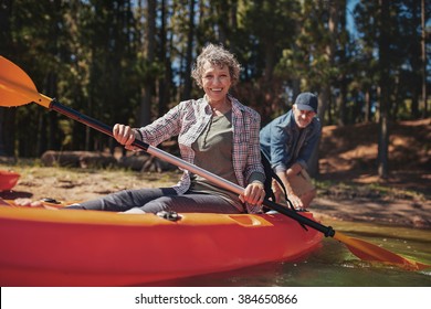 Portrait of happy senior woman in a kayak holding paddles. Woman canoeing with man in background on the lake. - Powered by Shutterstock